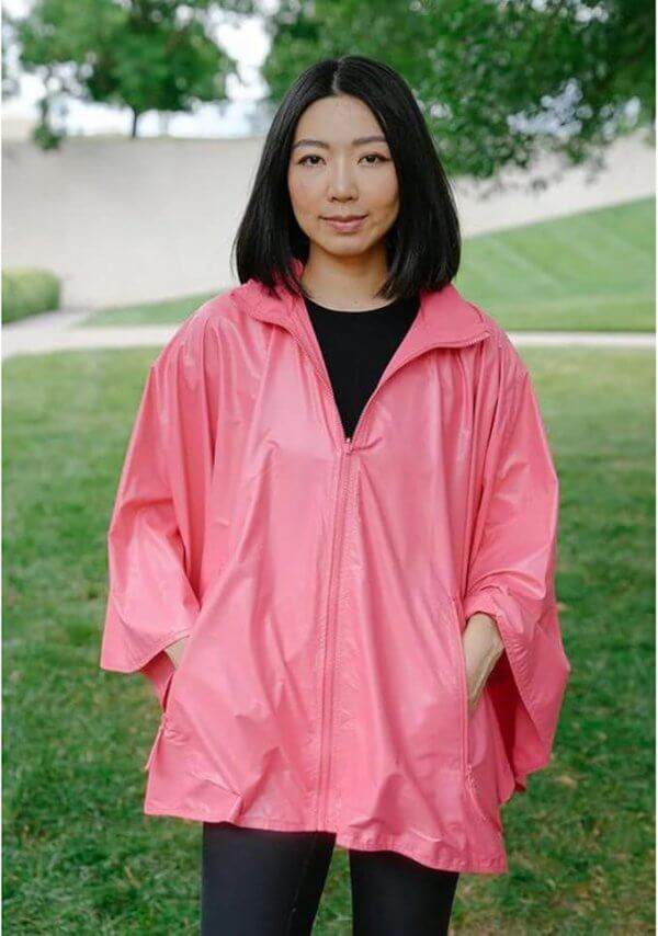 A person with shoulder-length hair wearing a bright pink poncho stands on a grassy area, surrounded by trees blossoming.