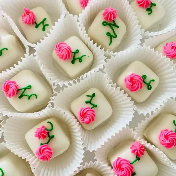 Square white petit fours with pink rose and green leaf icing designs, each nestled in a white paper cup.