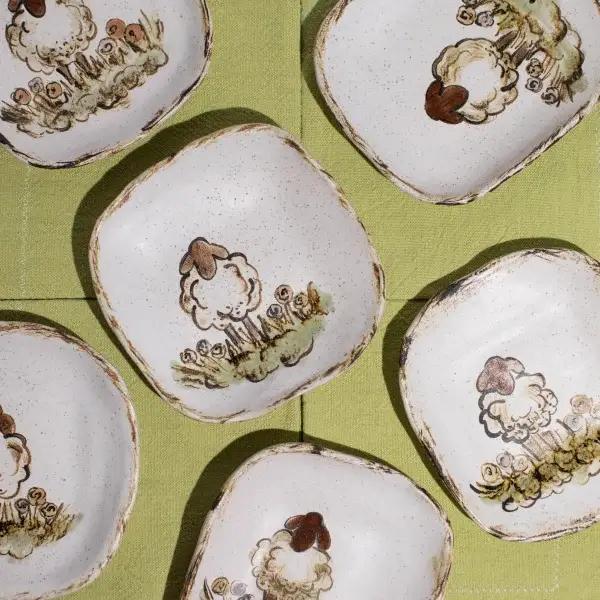 Six square ceramic plates featuring a charming sheep design, beautifully arranged in an overlapping pattern on a lush green fabric background.