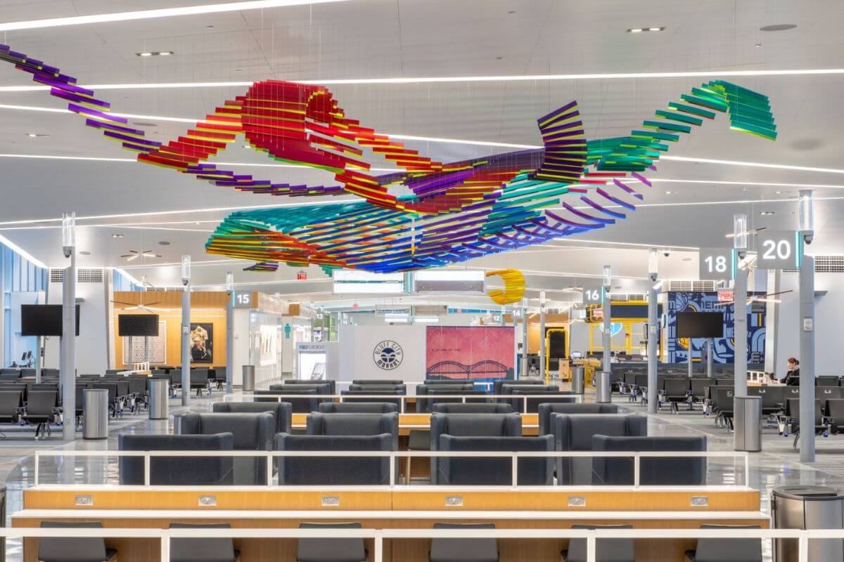 Spacious airport terminal with modern seating, digital screens, and a colorful abstract art installation suspended from the ceiling. Gate numbers 15 to 20 are visible, serving passengers on direct flights from Memphis.