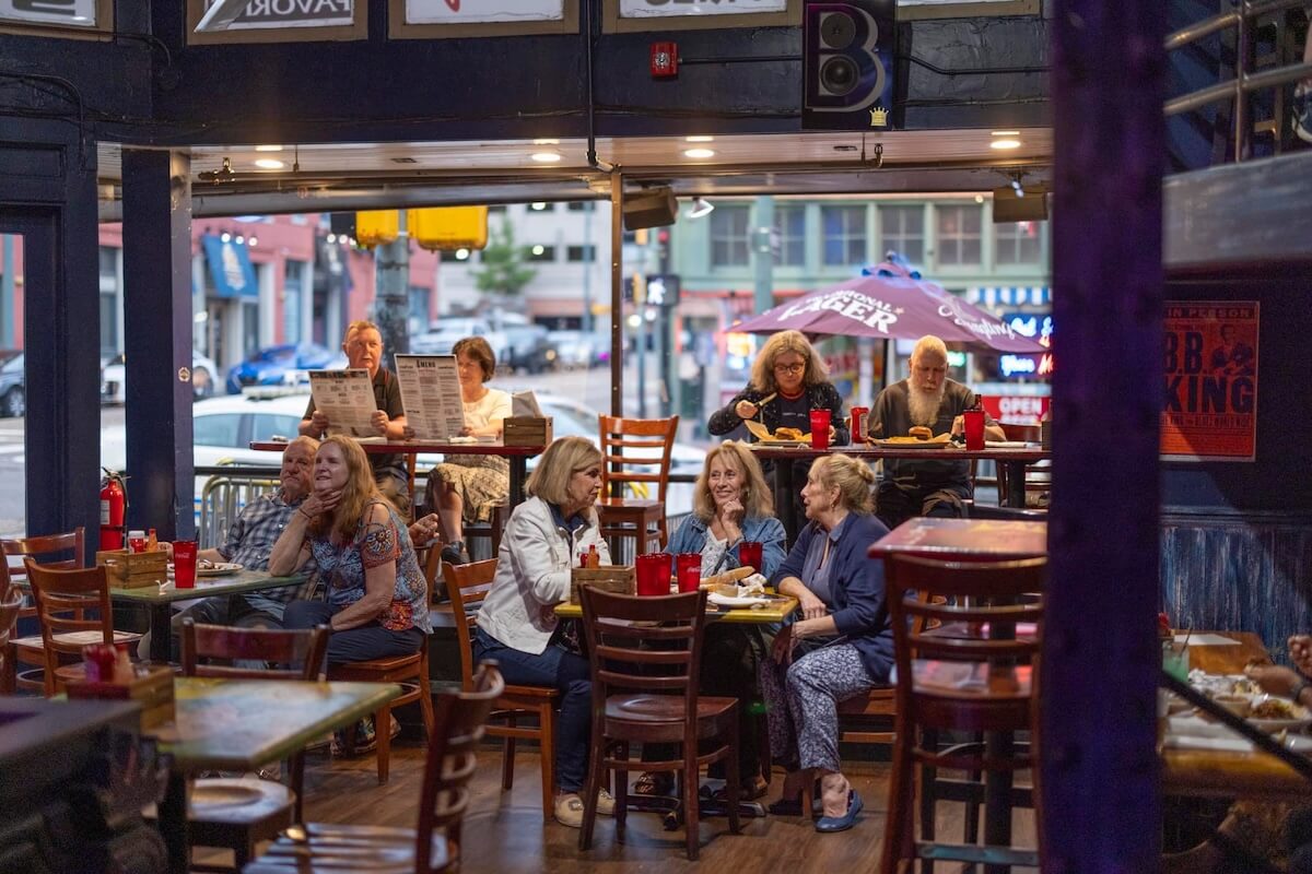 People seated at tables in a bustling Memphis restaurant with large windows. The background features umbrellas, street views, and a mix of diners eating and talking, embodying the vibrant essence of this city's dining scene.