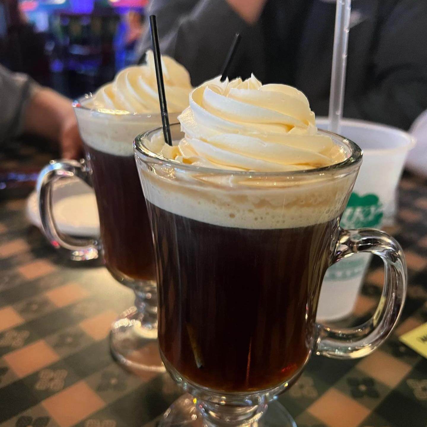 Two glass mugs of coffee topped with whipped cream and stirrers sit on a checkered table, creating the perfect cozy ambiance for your Memphis dining experience.