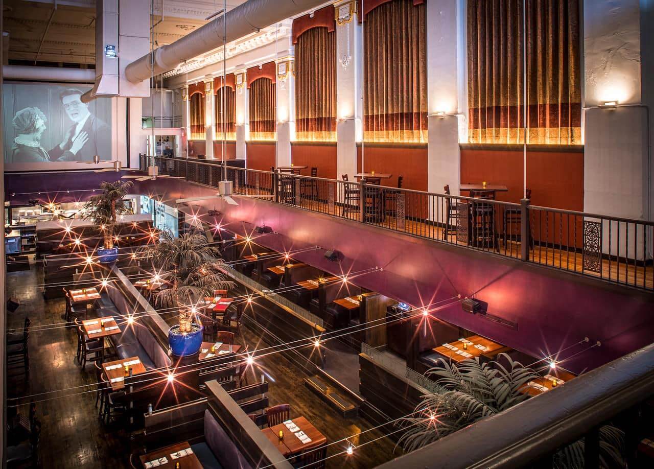 Spacious, dimly lit dining area with tables and lights sets the scene in this Memphis gem. Large curtains and a vintage movie screen adorn the walls, while an elevated seating area and potted plants guide the eye throughout the inviting restaurant interior.