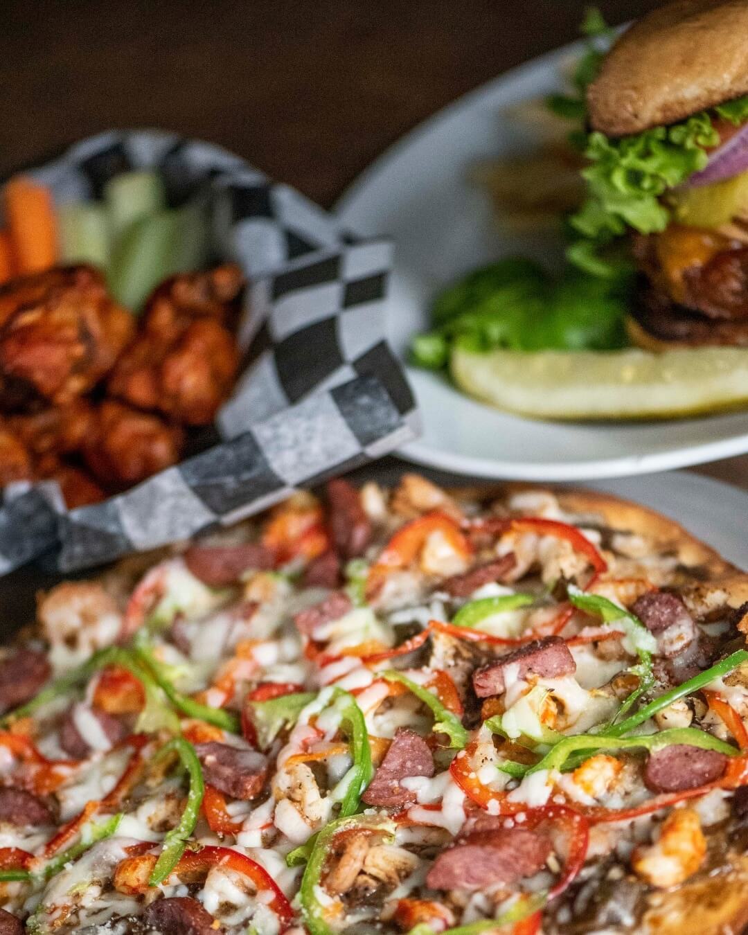 A close-up of a pizza topped with peppers and meats is the star, flanked by a basket of wings and fresh vegetables. In the background, a burger with fries and a pickle awaits, offering just a taste of what Downtown Memphis dining has to offer.