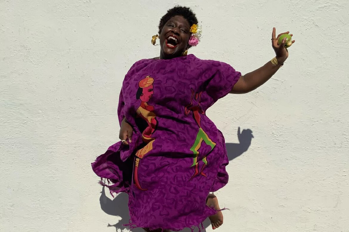 A person in a vibrant purple dress with a colorful design, joyfully jumps against a plain white background, channeling a Hatshepsut-like elegance while holding a green apple and wearing flowers in their hair, embodying the spirit of Bandele's artful FACES.