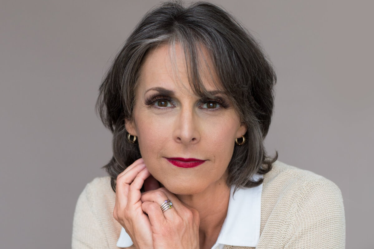 Stacey Saed, with her short gray hair and striking red lipstick, gazes directly at the camera. Clad in a light-colored sweater over a white collared shirt, she rests her hands thoughtfully near her chin.