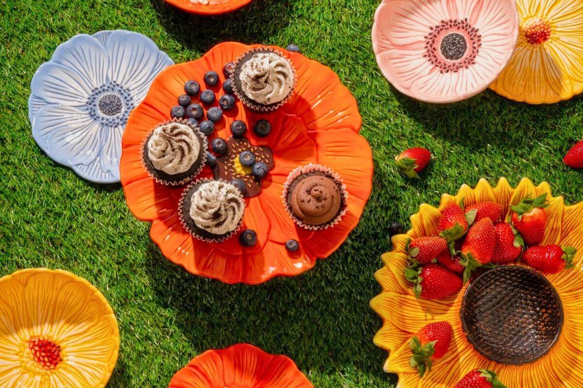 Colorful flower-shaped plates on grass, holding cupcakes, blueberries, and strawberries, create a vibrant picnic scene perfect for showcasing Memphis FINDS this March.