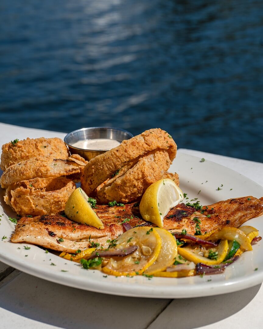Grilled fish with lemon wedges, grilled onions, and fried onion rings on a white plate by the Mississippi waters, offers a delightful stop along your Culinary Trails journey.