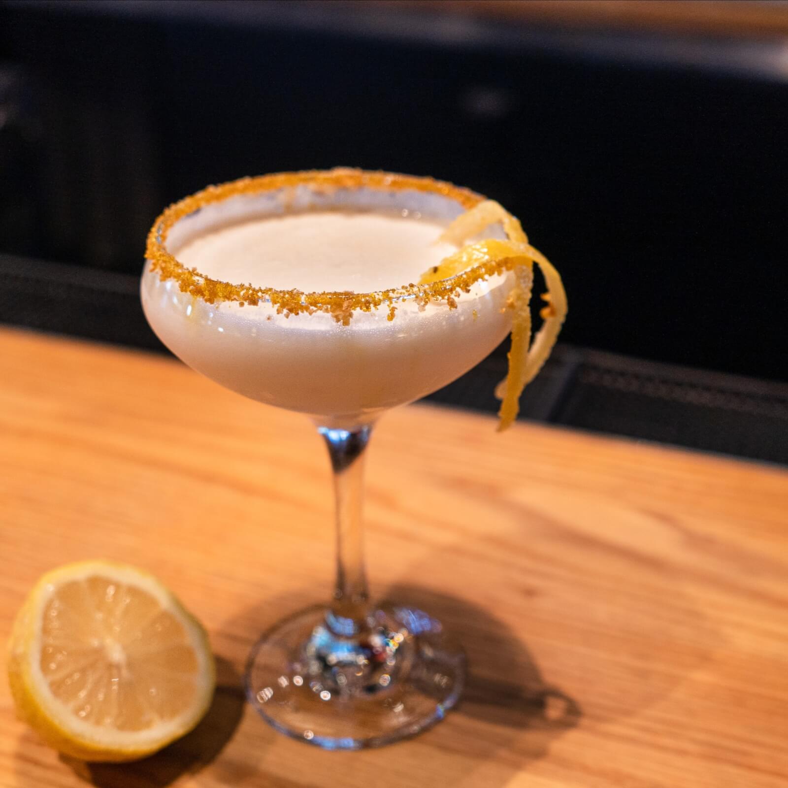 A cocktail in a coupe glass with a sugared rim and lemon twist garnish sits on a wooden surface next to a halved lemon, reminiscent of Southern charm along the culinary trails of Mississippi.