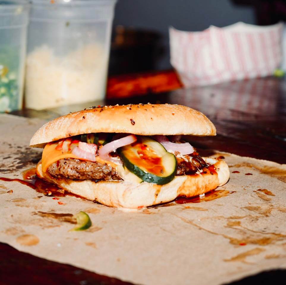 A burger with cucumbers, onions, and sauce on a seeded bun sits on a sheet of brown paper, evoking the delicious discoveries found along Mississippi's Culinary Trails. In the background, two plastic containers await their turn.