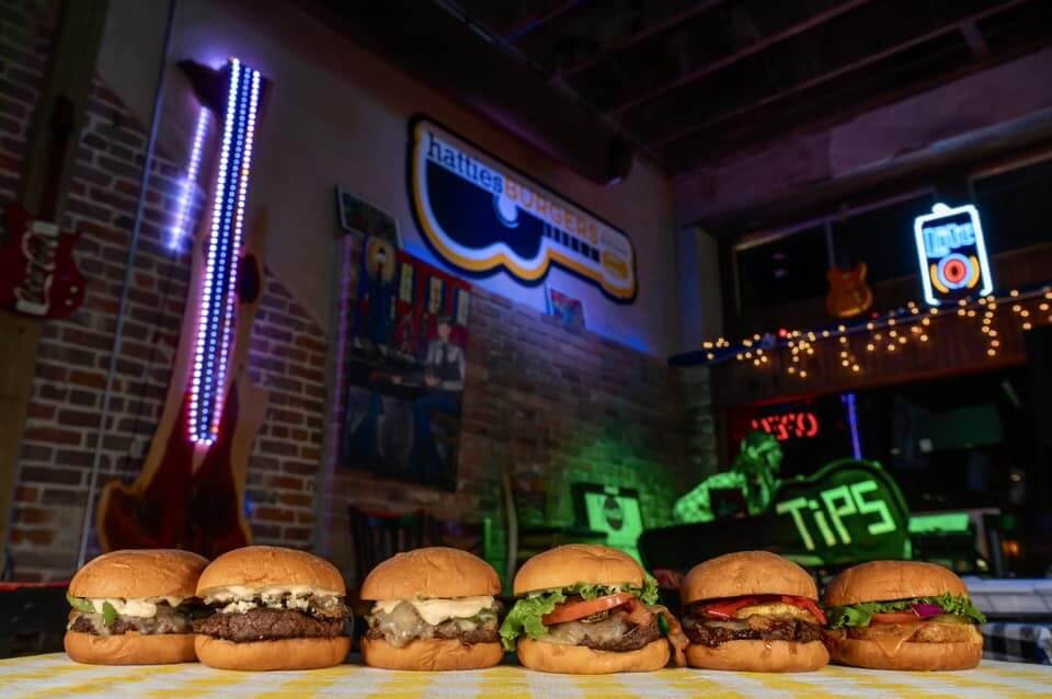Five burgers lined up on a checkered table in a dimly lit Mississippi restaurant, where neon signs glow and a guitar graces the wall.