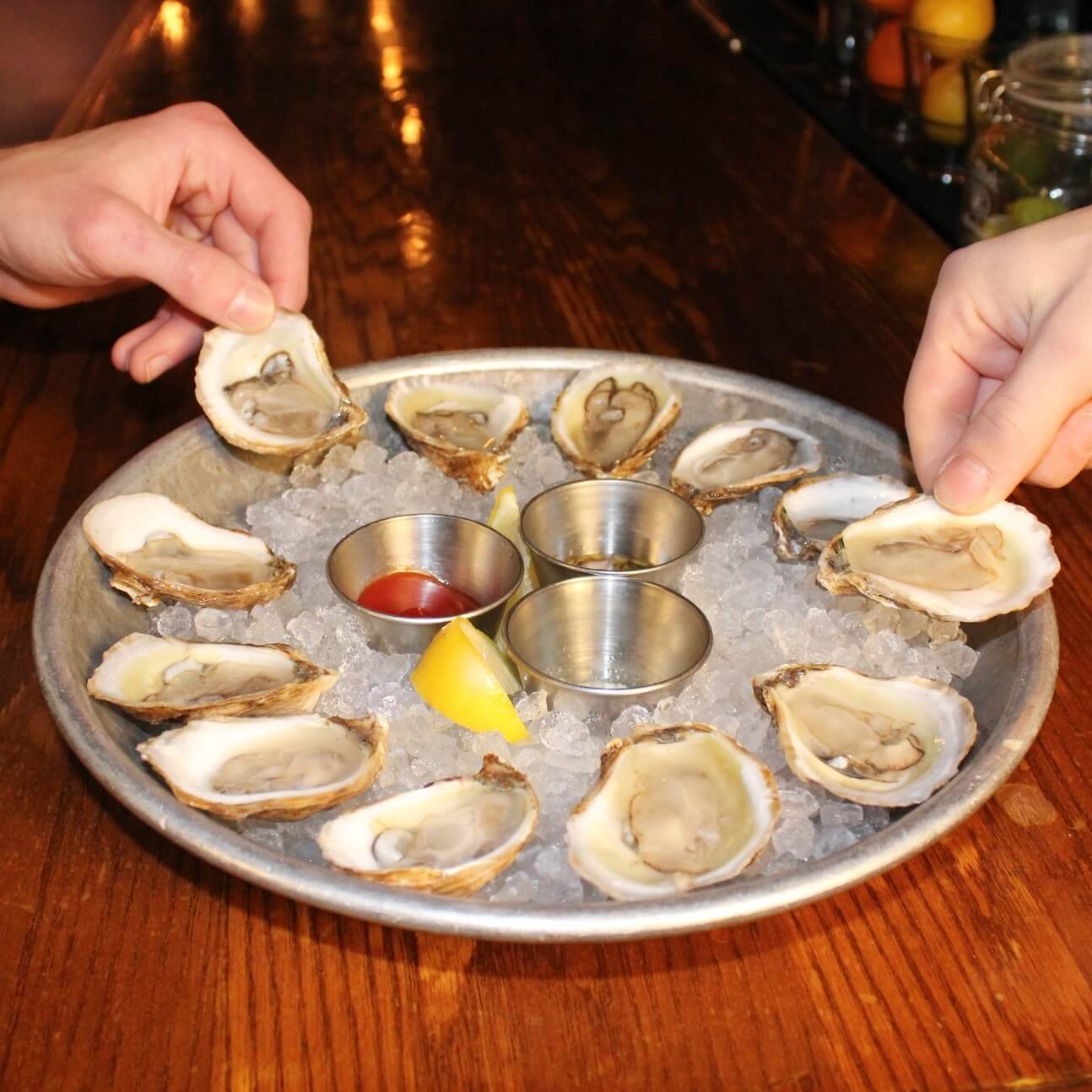 Two hands reach for oysters on a platter of crushed ice, surrounded by lemon wedges and dipping sauces.