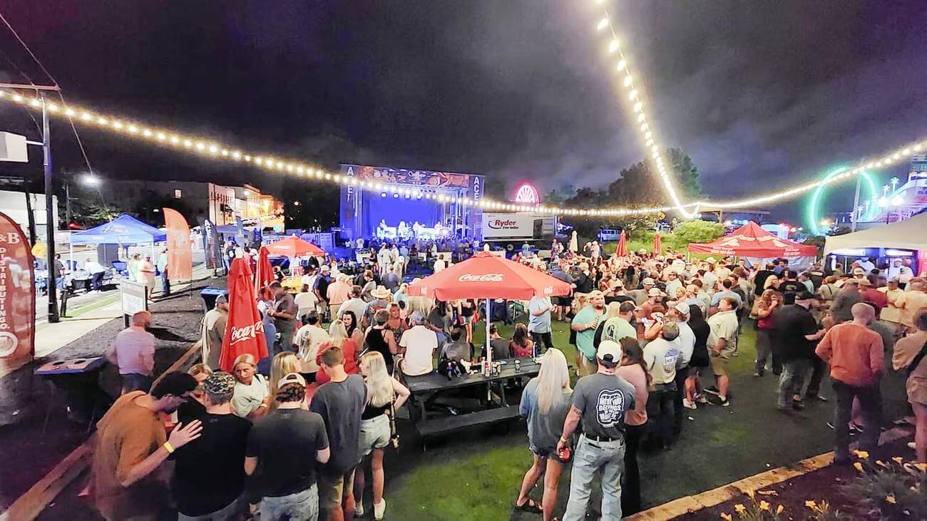 Outdoor night concert with a crowd under string lights. People gather around tables and a stage with musicians, enjoying the vibrant Mississippi vibe. Red umbrellas and vendor tents add charm as the aroma of culinary delights fills the air, enhancing the festive atmosphere.