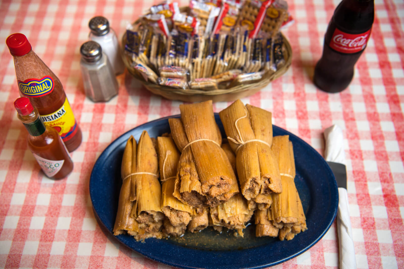 A plate of tamales on a checkered tablecloth, surrounded by hot sauce bottles, salt and pepper shakers, a basket of crackers, and a refreshing bottle of Coca-Cola.