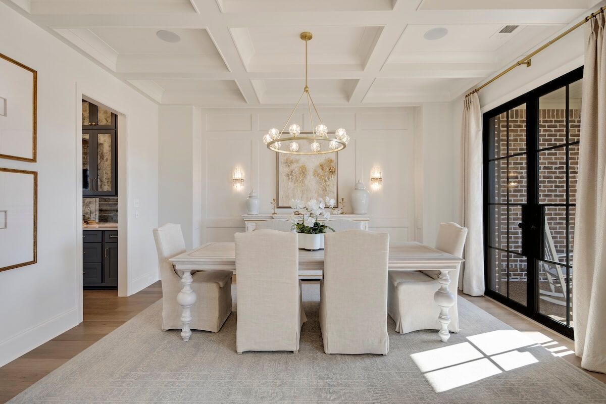 Elegant dining room inspired by Magnolia Homes, featuring a rectangular table, six beige upholstered chairs, a chandelier, and a large window with curtains. The white walls are adorned with decorative molding and framed art.