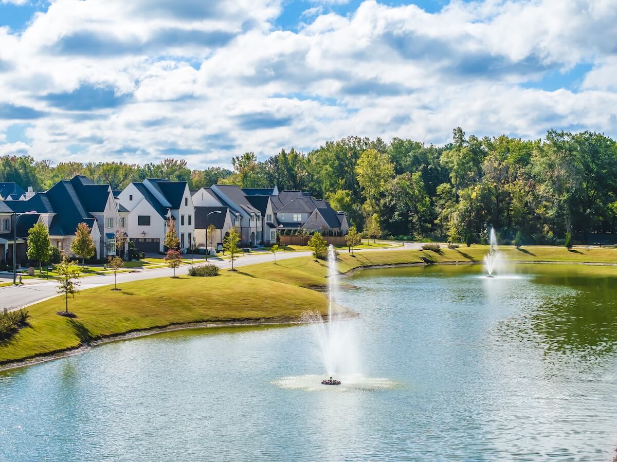 A suburban housing development featuring Magnolia Homes with several modern designs surrounds a picturesque pond and two fountains on a sunny day. Tall trees and lush grassy areas embrace the serene waters, crafting a peaceful community.