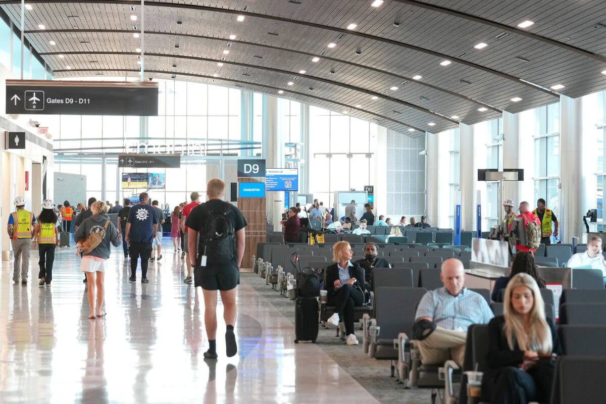 People sit and walk in a busy airport terminal near gate D9. Some travelers check their devices for updates on direct flights from Nashville, while others wait or move through the area. Signs and large windows are visible.
