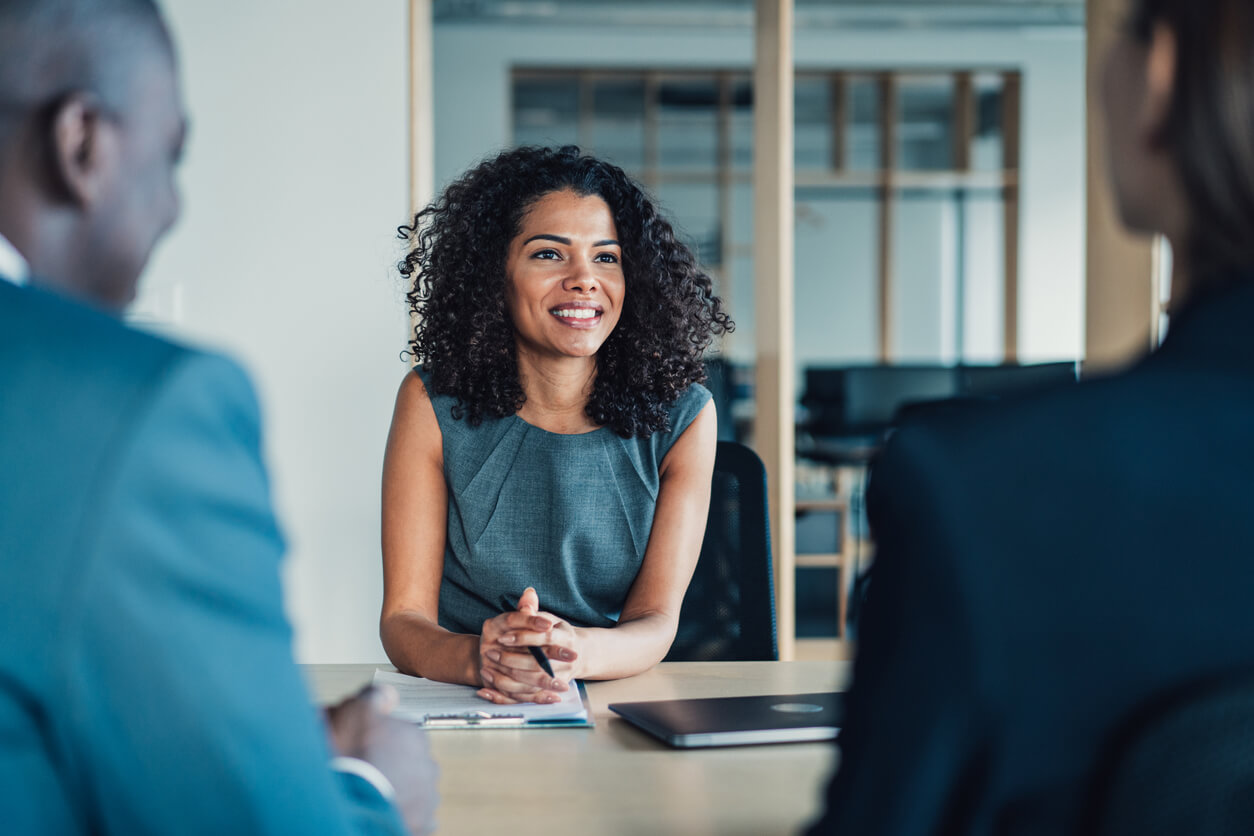 A woman with curly hair sits at a table, smiling, during a meeting about the collaborative divorce process with two other people. A laptop and a notepad are on the table.