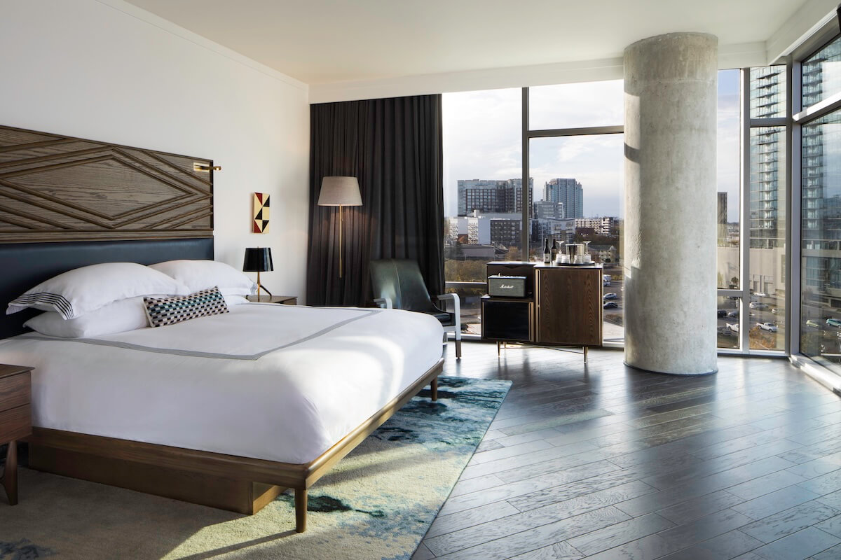 Modern hotel room with a large bed, floor-to-ceiling windows, a chair, and a small amplifier. Enjoy the Nashville cityscape's tall buildings under a partly cloudy sky.