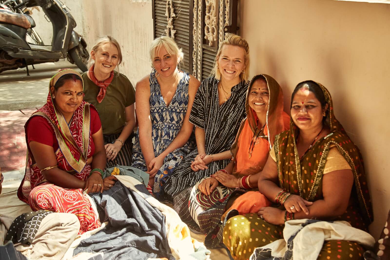 Six women, some in traditional attire, sit together on the ground, exuding warmth and joy as they smile at the camera. A scooter leans against a wall behind them.