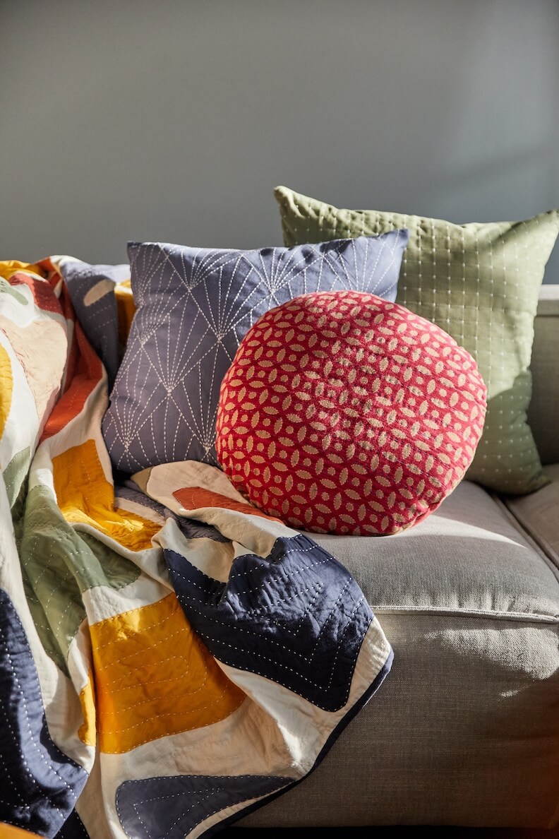 This cozy couch setup speaks to design and collaboration, featuring a patterned quilt, two square pillows in shades of blue and green, and a round red pillow adorned with white patterns.