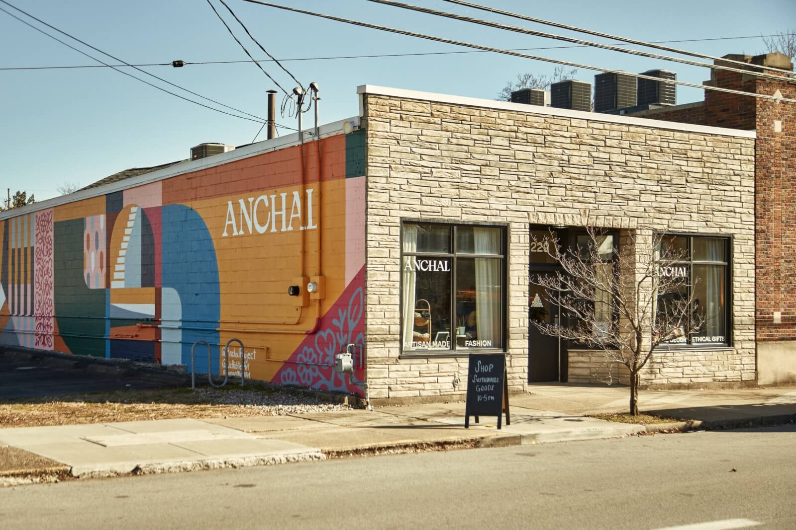 Street view of a store with a colorful mural on its side wall, featuring geometric shapes. The facade displays "ANCHAL" and a sidewalk chalkboard sign stands outside. This Southern nonprofit is committed to empowering marginalized women through opportunities in design and culinary arts.