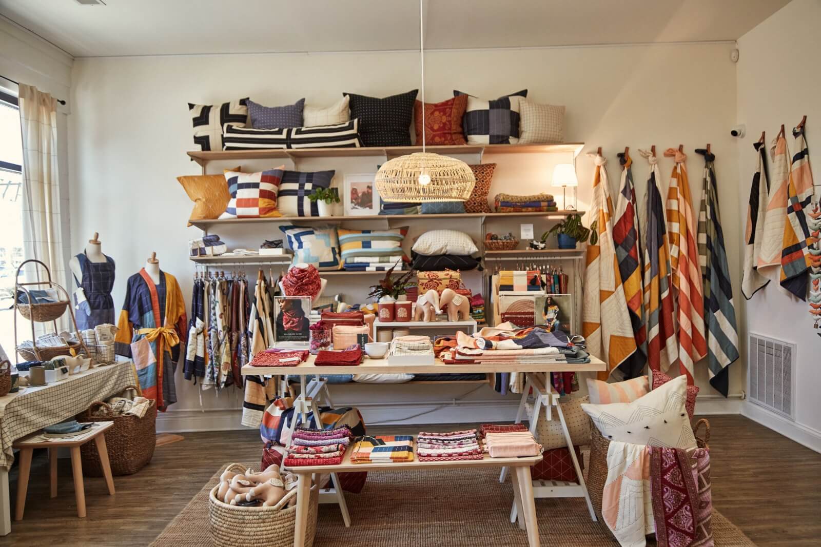 Store interior features shelves lined with cushions, a display of colorful textiles, and a table brimming with folded fabrics.