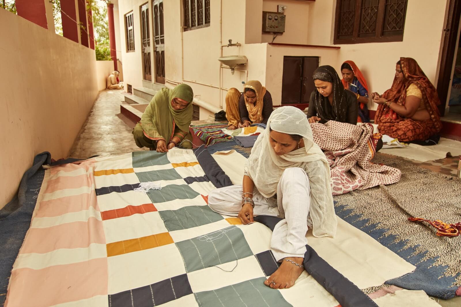 Women in traditional attire sit on the ground, collaborating on a large, colorful quilt outside a building. 