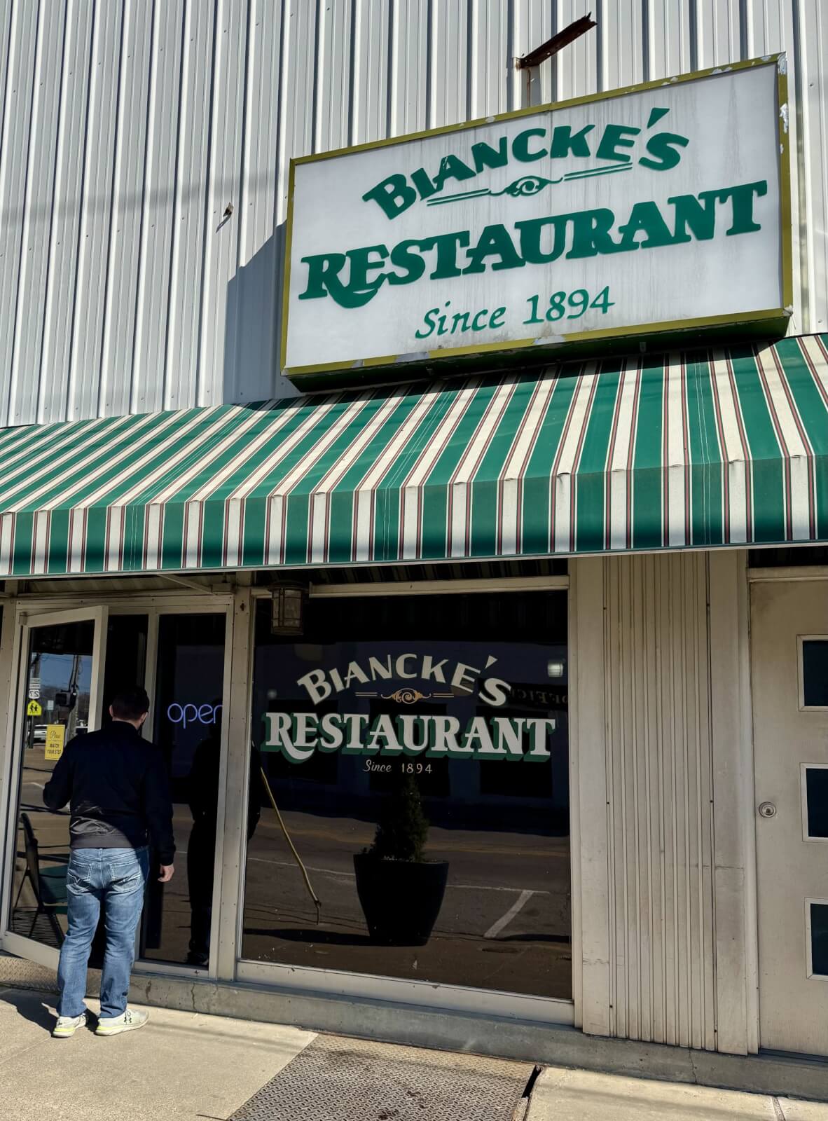 A person stands outside Biancke's Restaurant in Cynthiana, Kentucky, established in 1894. The building features a green-striped awning and a large sign proudly displaying the restaurant's name.