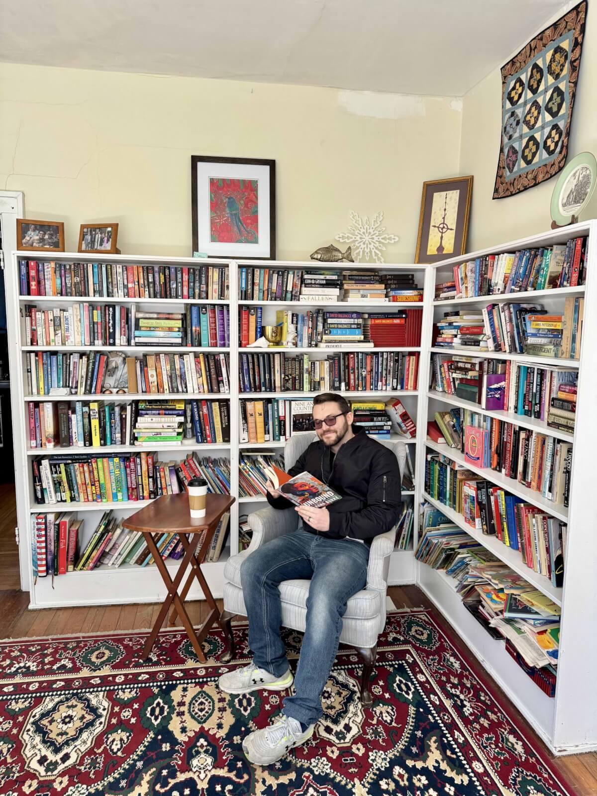 A person sits in a chair, reading a book in a cozy room reminiscent of Cynthiana, Kentucky. Two white bookshelves brim with books while a small wooden table with a cup rests nearby. A patterned rug adds charm to the space.