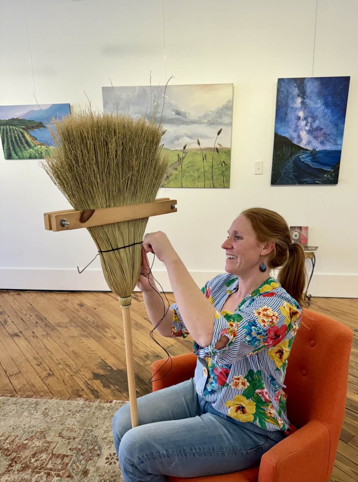 In her cozy Cynthiana, Kentucky home, a woman in a floral shirt sits on an orange chair, meticulously crafting a broom made of twigs. Artworks hang on the wall behind her.