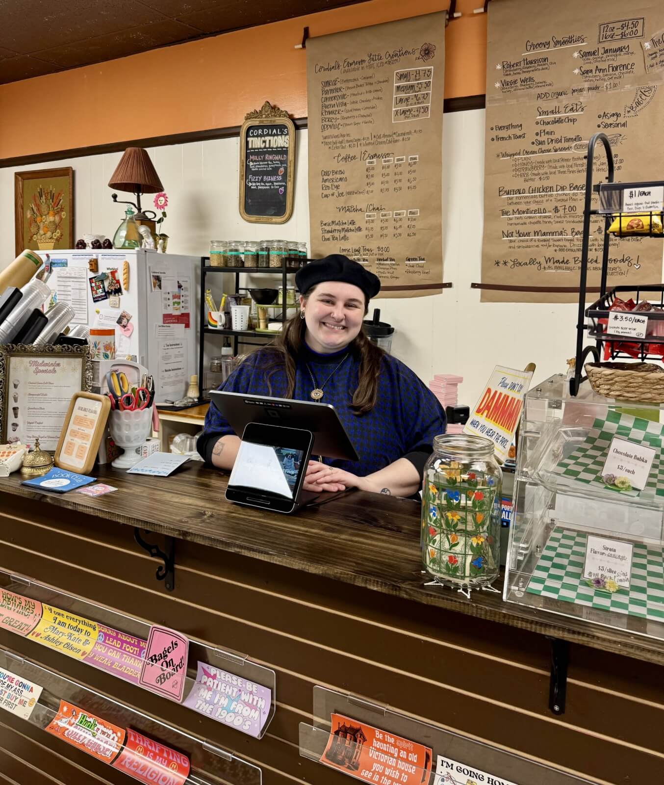 A person in a beret stands behind a cafe counter with an iPad in charming Cynthiana, KY. Colorful jars and candies adorn the counter, while menus grace the wall behind, capturing Cynthiana's vibrant spirit.