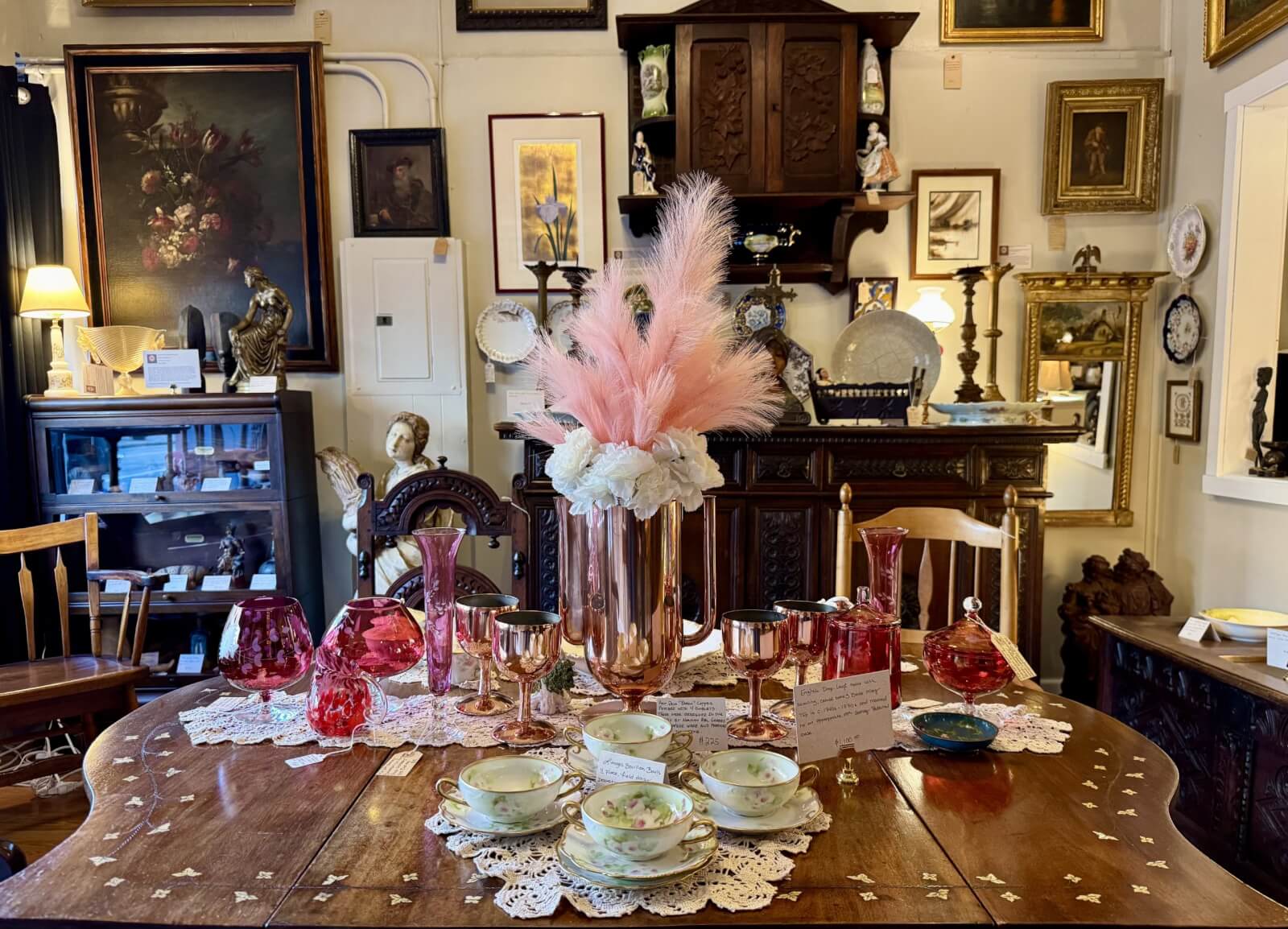 An antique store display in Cynthiana, Kentucky, features a wooden table adorned with vintage tea cups, pink and clear glassware, and a centerpiece of pink feathers. The surrounding walls showcase an array of framed artwork and decor, capturing the charm of Cynthiana.