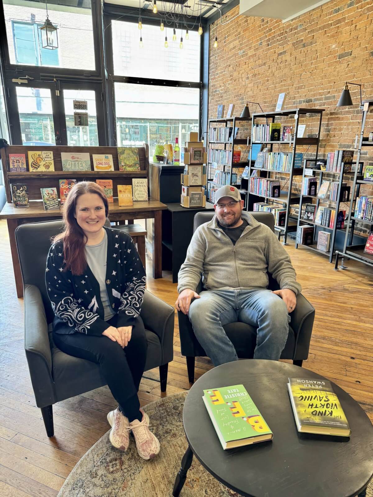 Two people are sitting in chairs at a cozy bookstore in Cynthiana, Kentucky. Books are displayed on shelves and tables around them, with a charming brick wall and large windows adding to the inviting ambiance.