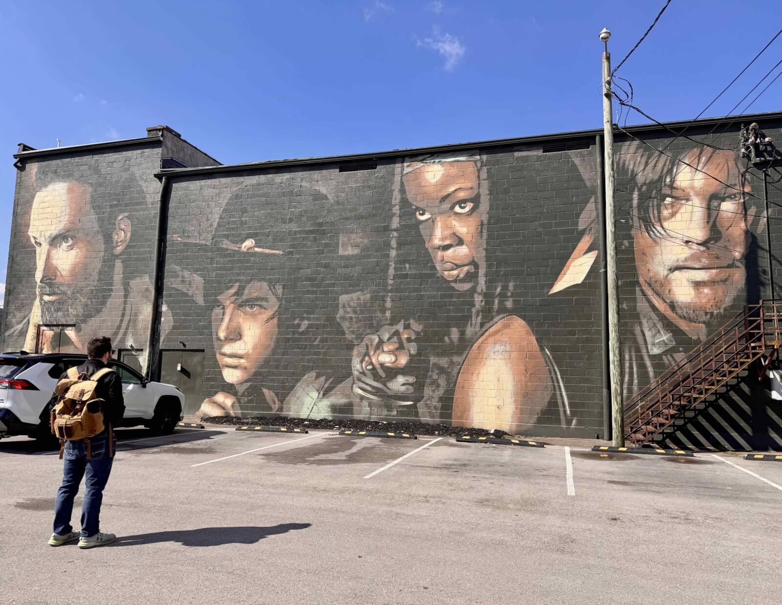 A person with a backpack stands in a parking lot in Cynthiana, Kentucky, admiring a large mural of four individuals on a building wall.