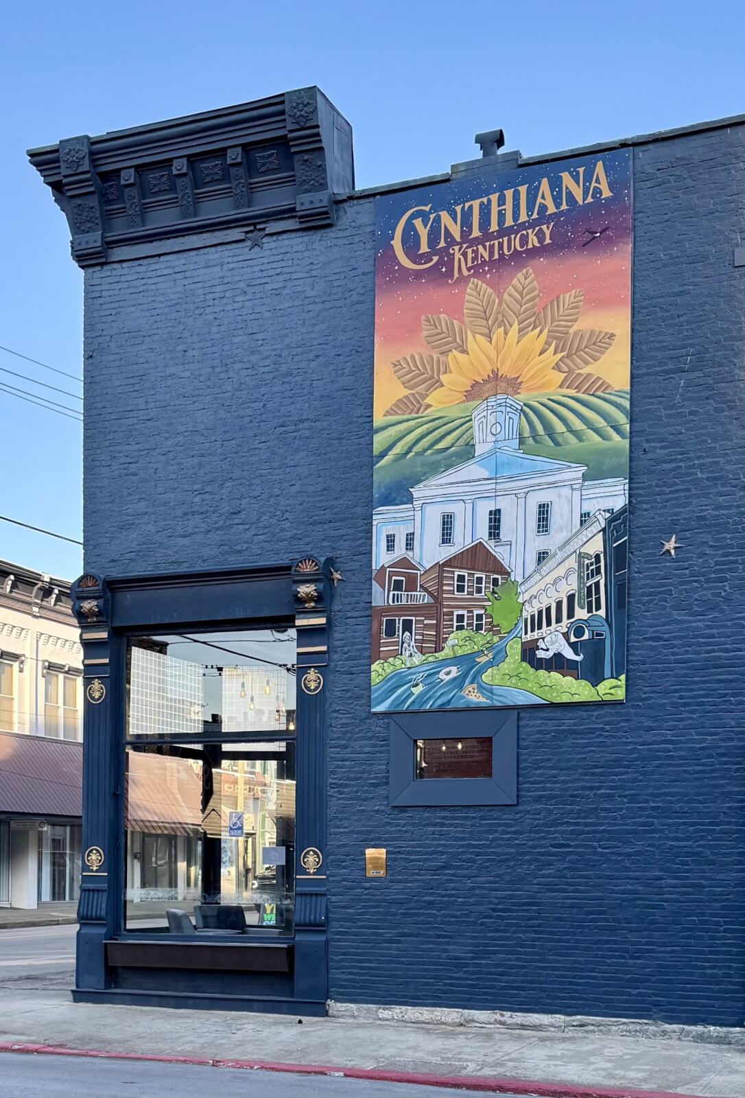 A blue building facade in Cynthiana, Kentucky, showcases a mural labeled "Cynthiana, KY," adorned with captivating landscape and architectural elements.