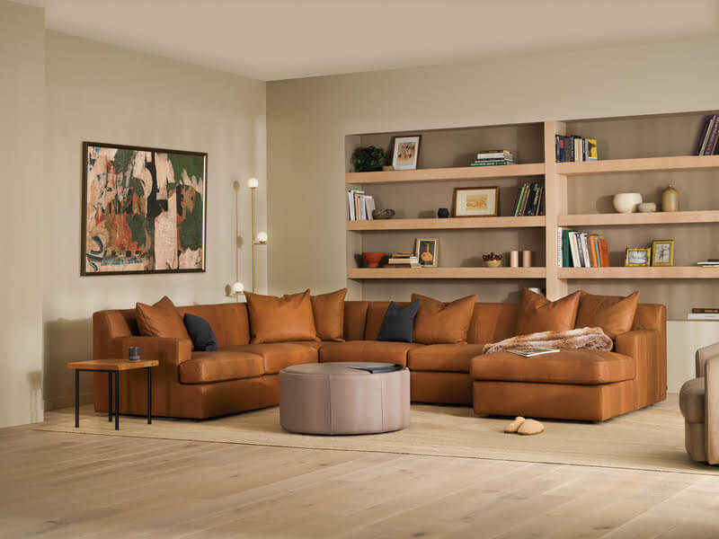 A living room inspired by the best home and gift stores in Birmingham, featuring a brown leather sectional sofa, a round ottoman, a side table, wall art, built-in shelves with books and decor on light wood flooring.