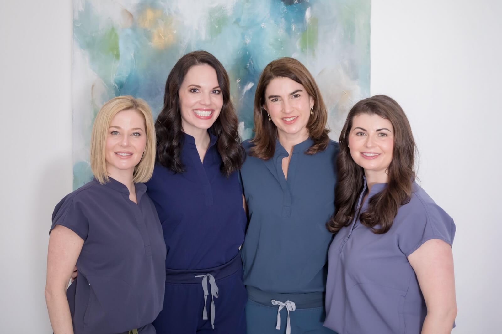 Four women wearing matching blue and purple scrubs stand together, smiling, in front of an abstract painting—ready to provide expert facials and skin services.