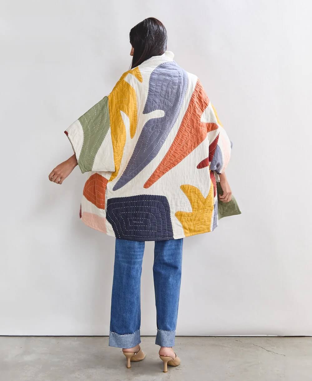 The individual, cloaked in an anchal-inspired colorful abstract-patterned quilted coat, blue jeans, and tan heels, stands with their back to the camera against a plain white background.