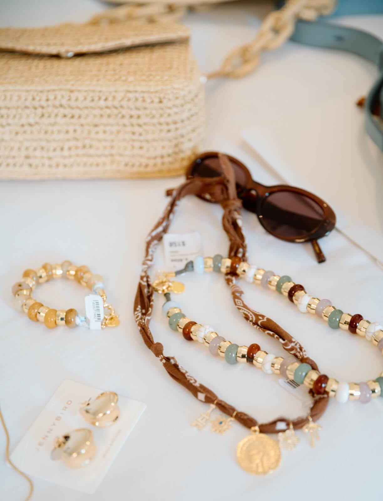 Polished & Playful: What to Wear This Spring - 9 A curated collection of assorted jewelry and accessories is artfully displayed, featuring a beaded bracelet, sunglasses, necklaces, and a woven bag. Perfect for WTW looks in Spring 2025.