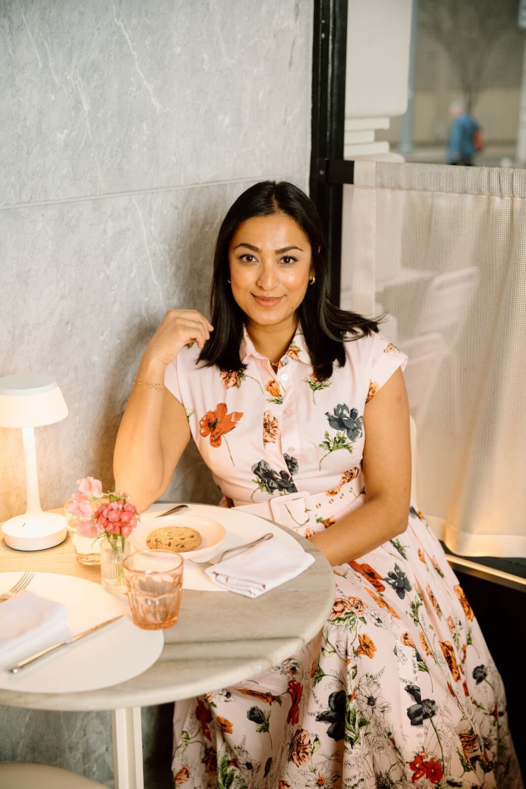 Polished & Playful: What to Wear This Spring - 3 A woman in a floral dress sits at a small, round table with a cookie on a plate and a glass of water. Celebrating Spring 2025, the scene is illuminated by a small lamp and features pink blossoms in a vase.
