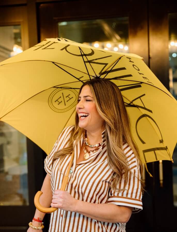 Polished & Playful: What to Wear This Spring - 10 A woman beams with joy, clutching a sunny yellow umbrella adorned with text. She complements her look with a striped shirt and elegant necklace, standing confidently in front of a dark door.