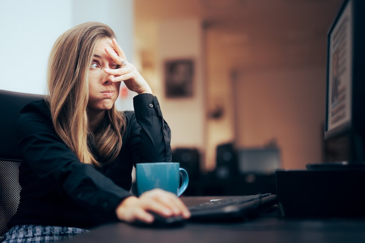 A woman sits at a desk with a computer, looking stressed with her hand on her face, contemplating phrases to retire from her workload. A blue mug is beside her.