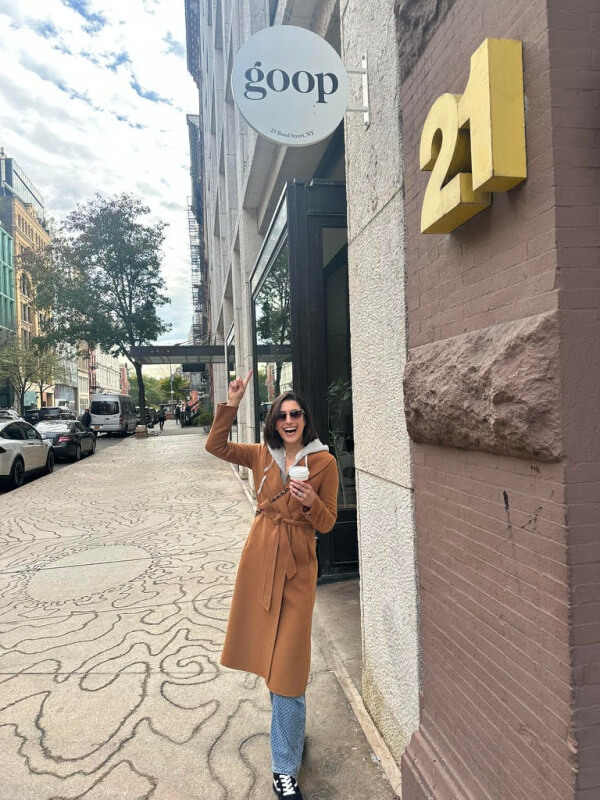 Outside the "goop" store, Jackie Laurian Long stands on the bustling city street in a brown coat, drink in hand, waving cheerfully amidst passing cars and swaying trees.