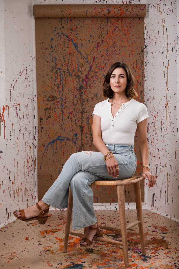 Jackie Long sits gracefully on a wooden stool, framed by a vibrant splatter paint backdrop. Her white top and denim jeans perfectly complement the lively colors surrounding her.