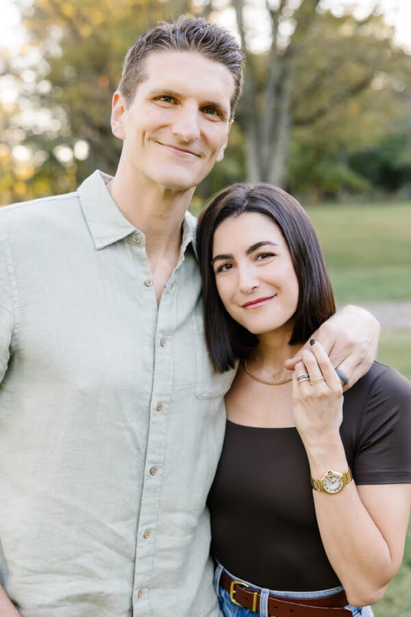Jackie and Jake stand closely together, both smiling warmly with trees in the background. Jackie rests her hand gently on Jake's shoulder, capturing a moment of connection and joy.