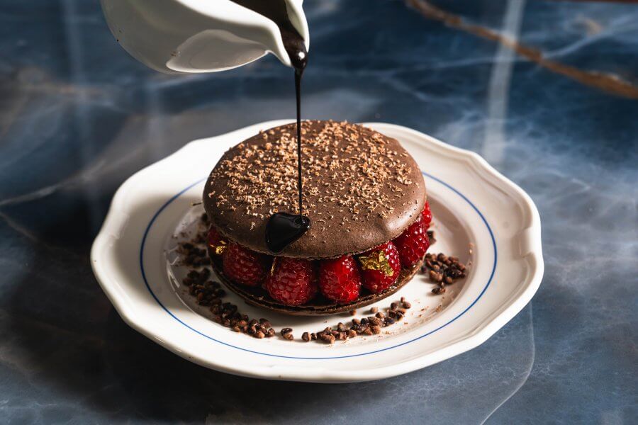 In one of Houston's top 10 new restaurants, a sumptuous dessert is adorned with glossy chocolate sauce, glistening atop a chocolate-covered top, accompanied by vibrant raspberries and elegant decorative elements on a pristine white plate.