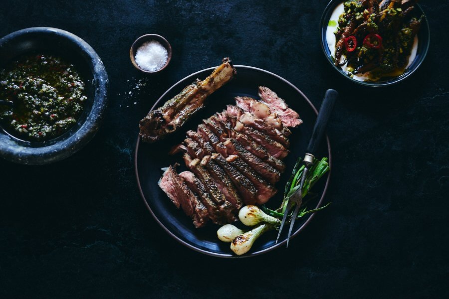 Sliced grilled steak with bone on a black plate, garnished with green onions, awaits you at one of the hottest new restaurants. Accompanied by a bowl of chimichurri sauce, a dish of peppers, and a small bowl of salt, this Houston restaurant promises an unforgettable dining experience.