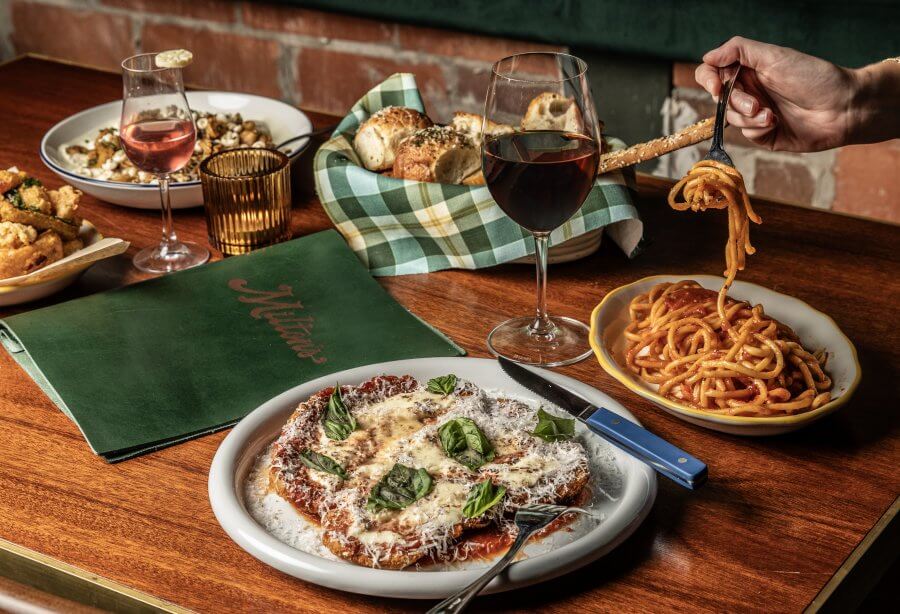 A table with plates of pasta, ravioli, bread, and two glasses of red wine awaits. A hand twirls spaghetti with a fork beside a green menu featuring "Milano." Discover the taste sensation at one of Houston's top new restaurants.