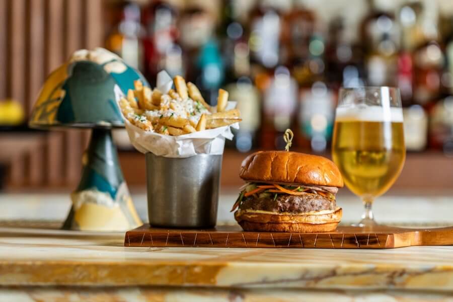 Burger on a wooden board with a side of fries in a metal cup and a glass of beer on a bar counter, capturing the essence of Houston's hottest restaurants.